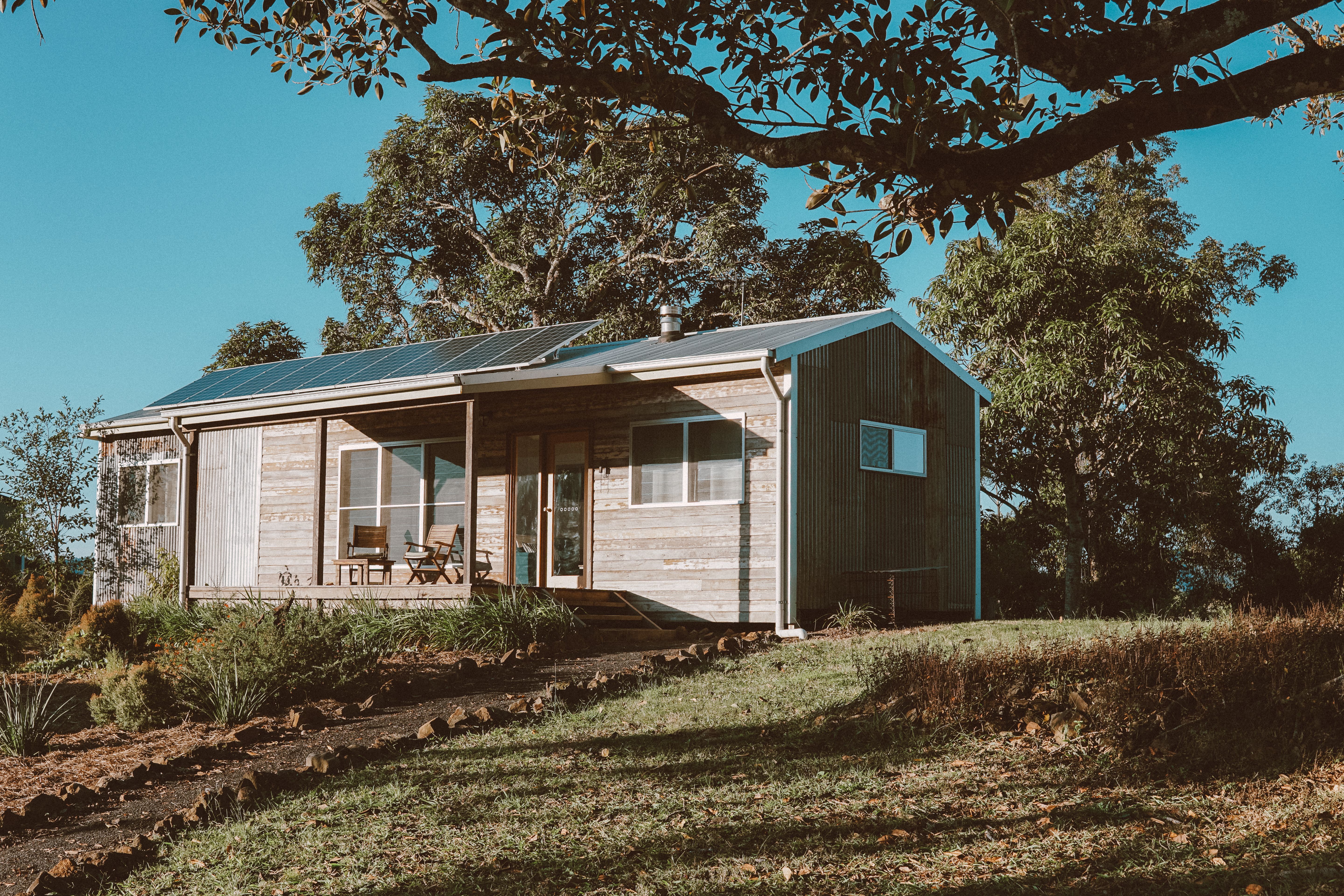 A bright sunny day with blue skies, a country house sits in the middle of the image with trees behind and a branch in front of the camera. A path leads up to the front door.