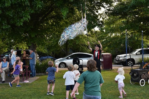 A man in a park holds up a bubble making device a group of children chase the bubbles below