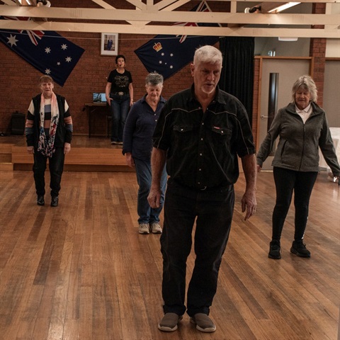 A community hall and four people are line dancing. Behind them there is an instructor on a stage. 