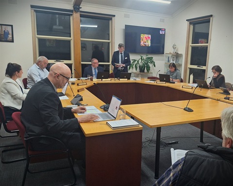The Pyrenees Shire Council Chambers featuring a horseshoe shaped table with councillors and staff seated around. There is one member of the public at the front of the room.