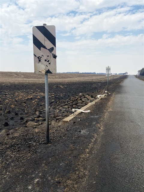A burnt road sign sits in the foreground next to a road that stretches into the distance. Beside the road are pastures burnt and black.