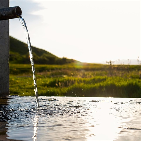 Water is flowing from a pipe into a container. In the background are fields and a setting sun.