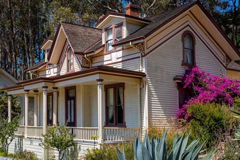 A two story country cottage with a front porch, and dormer windows on the second floor. There is a colourful garden around the house.