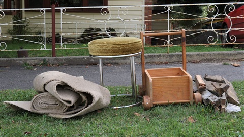 The grass verge in front of a house, hard rubbish including carpet, a stool, a timber drawer and a small pile of scrap wood. 