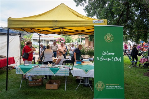 A stand at a market, The marquee has a yellow cover and tables with green tablecloths. There are people behind the table and several customers The sign next to it reads 'Welcome to Beaufort CWA'