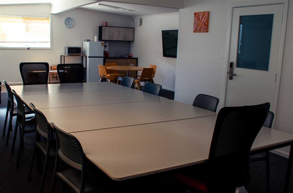 Room with large table and chairs, a window at the back of the room and a kitchenette to the side